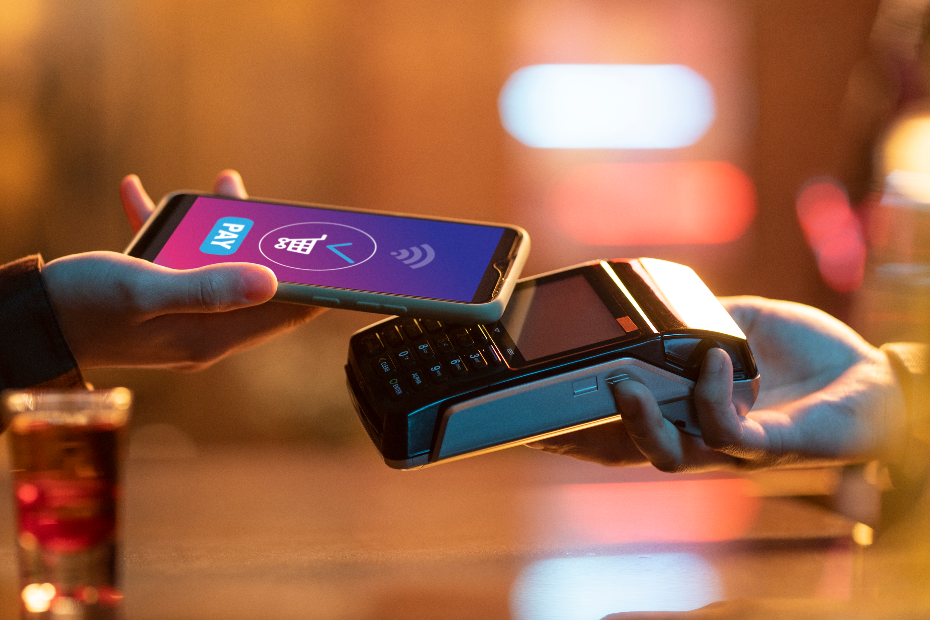The image depicts a hand holding a phone with a mobile wallet app open, hovering over a contactless payment terminal.